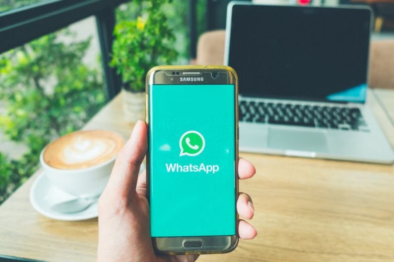 Smartfon marki Samsung z włączoną aplikacją WhatsApp na tle biurka, na którym stoi laptop i filiżanka kawy.