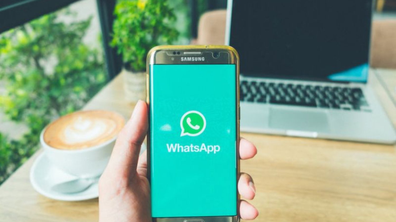 Smartfon marki Samsung z włączoną aplikacją WhatsApp na tle biurka, na którym stoi laptop i filiżanka kawy.