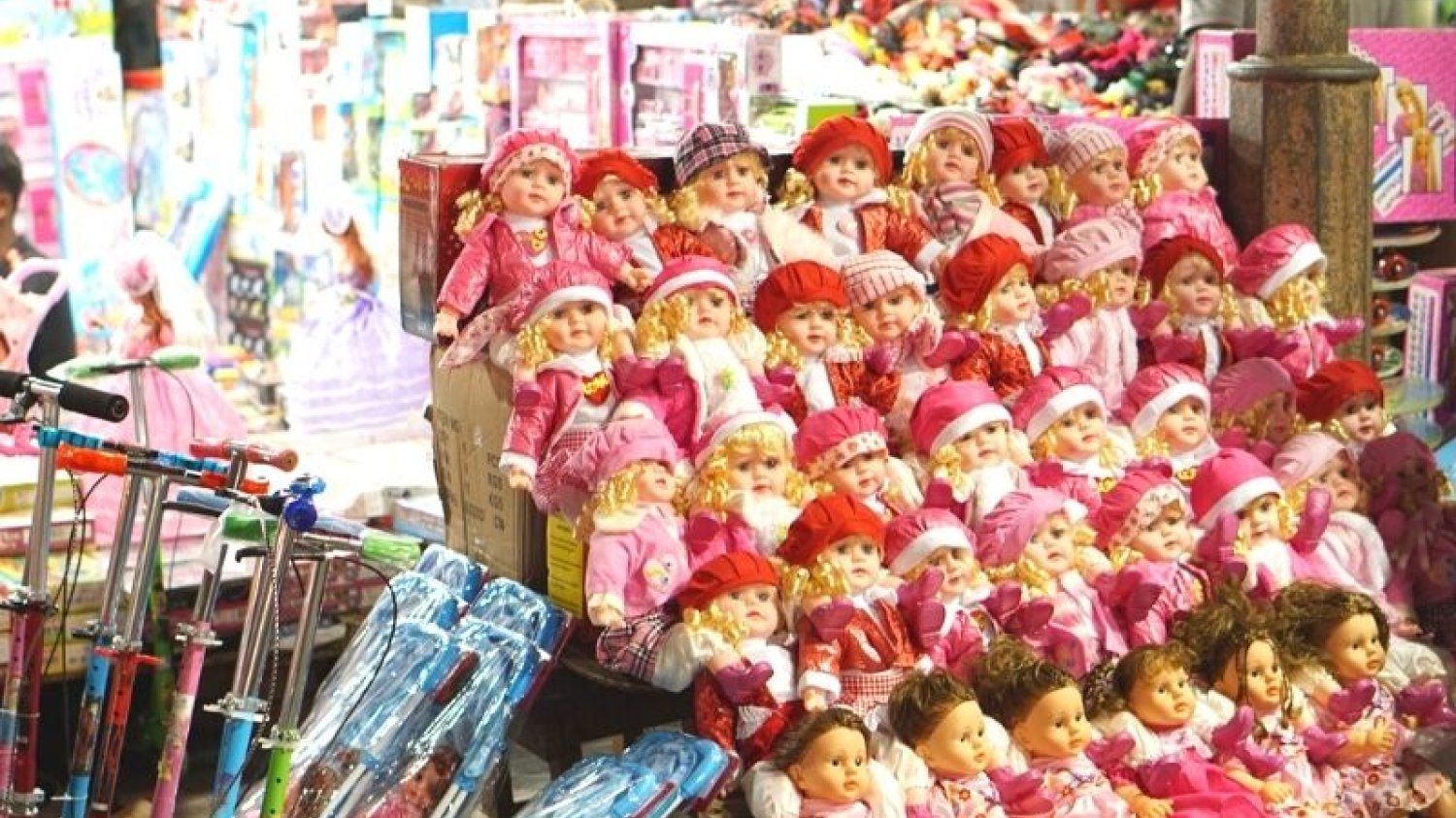 Lalki w różowych ubrankach na wystawie sklepowej, obok stoją kolorowe hulajnogi dla dzieci