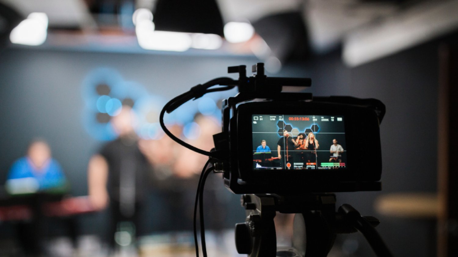 Studio filmowe: na pierwszym planie widzimy kamerę, która filmuje występ zespołu muzycznego. Zespół widzimy przez obiektyw kamery.
