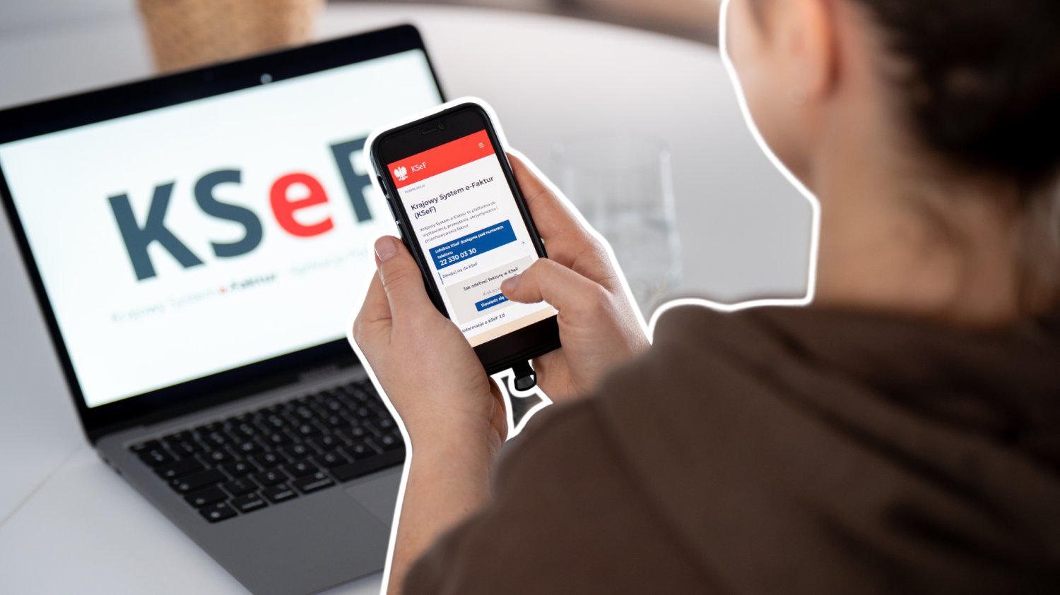 Pierwsze rozliczenie KSeF już 20 maja. Firmy mogą słono zapłacić za bałagan Kobieta trzymająca telefon z aplikacją KSeF. W tle laptop ze stroną KSeF.
