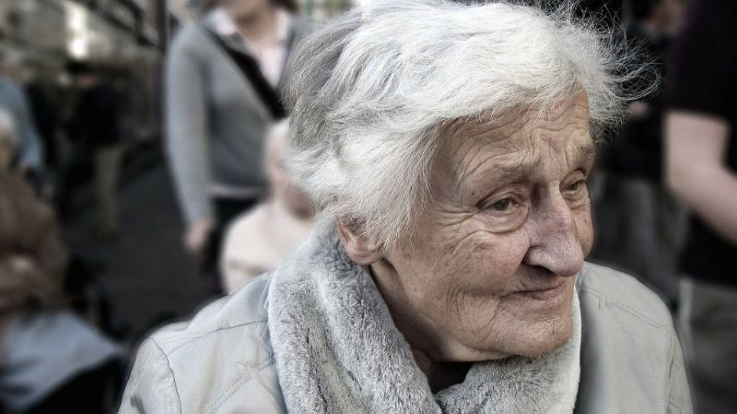 Starsza kobieta z nieszczęśliwą, zagubioną miną stoi wśród tłumu ludzi.