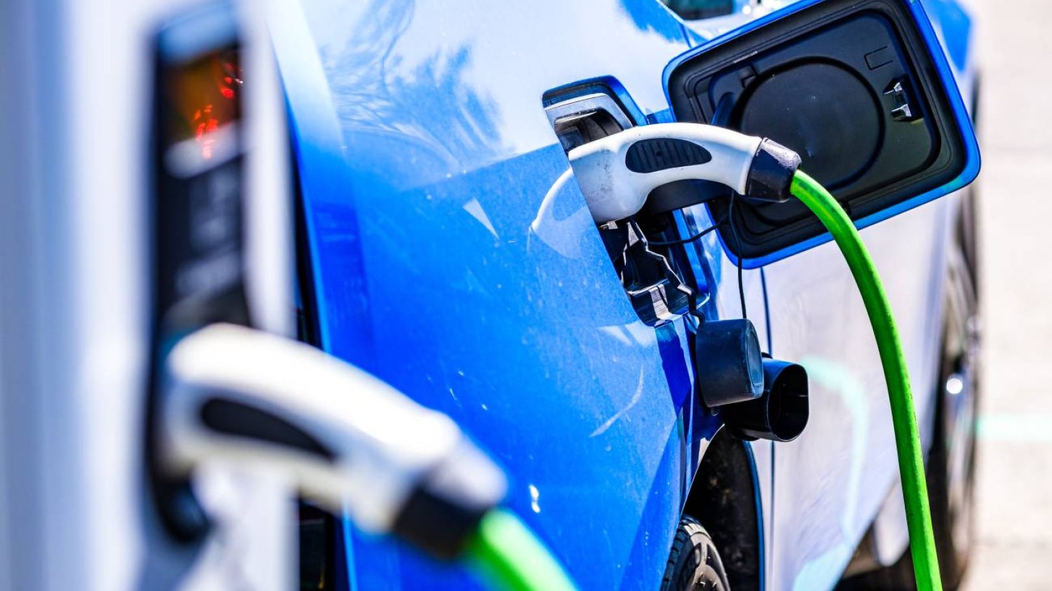 Niebieski samochód elektryczny przy stacji ładownia prądem. Zielona wtyczka ładuje pojazd.
