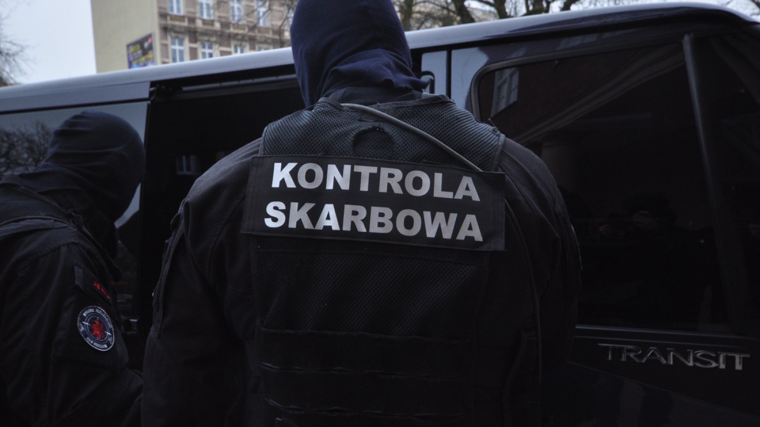 Policja skarbowa stoi przy wyjściu z ciemnego samochodu. Na pierwszym planie mężczyzna w kominiarce z napisem "kontrola skarbowa" na plecach.