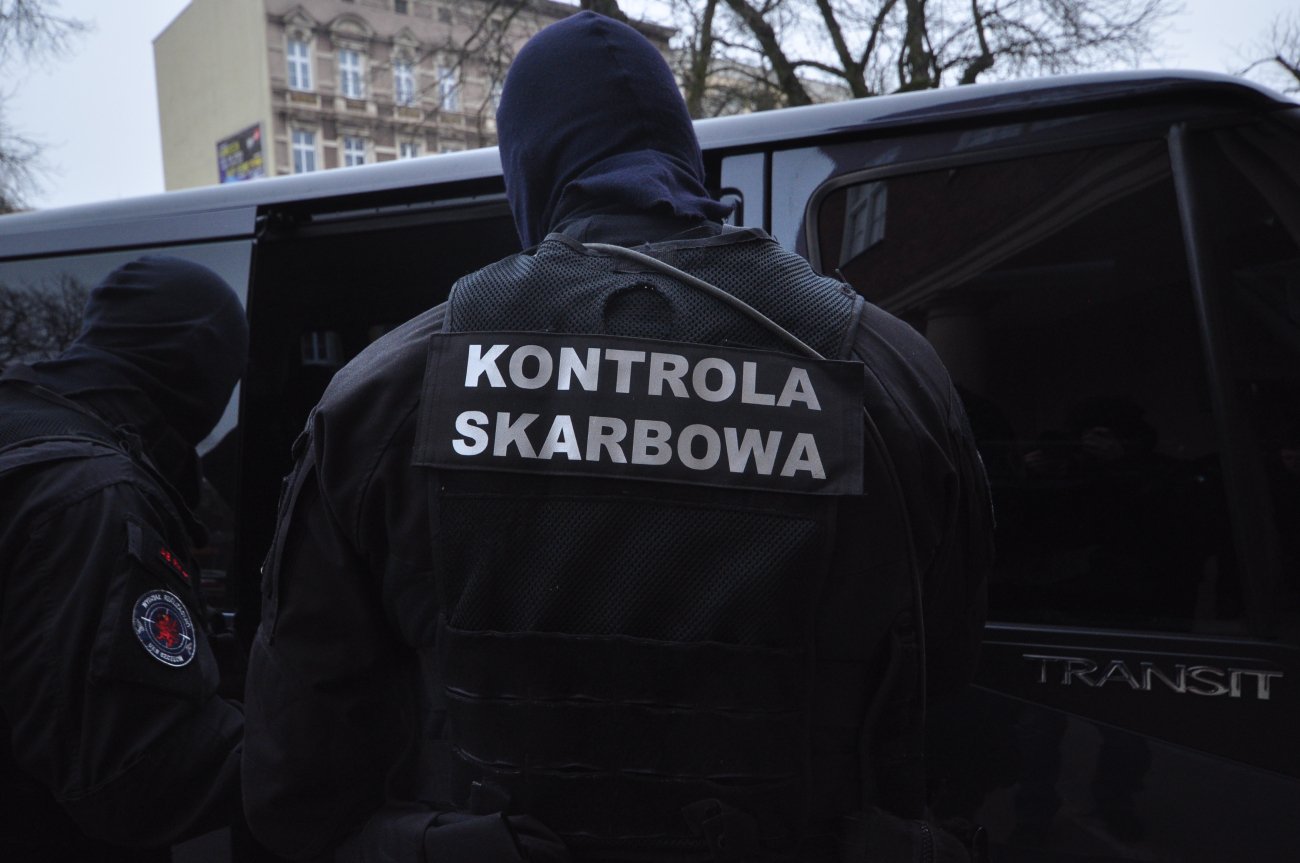 Policja skarbowa stoi przy wyjściu z ciemnego samochodu. Na pierwszym planie mężczyzna w kominiarce z napisem "kontrola skarbowa" na plecach.