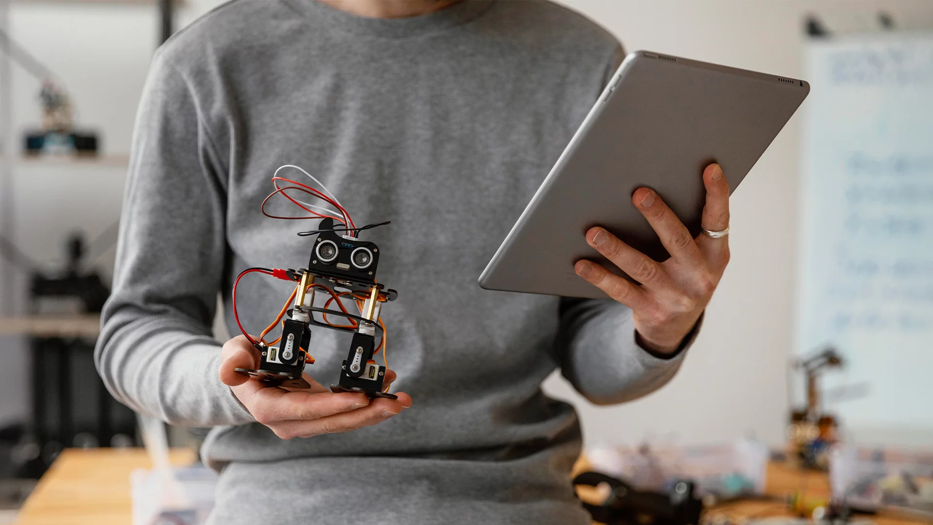 Osoba stoi przy biurku – w jednej ręce trzyma tablet, w drugiej małego robota. Holcim w ramach akcji Inżynierowie Jutra podpowiada, jak zatrzymać dobrego pracownika w Polsce.