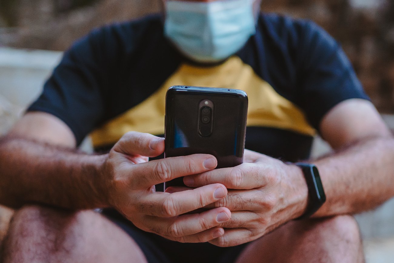 mężczyzna w maseczce siedzący na schodach z telefonem i mający na ręku opaskę