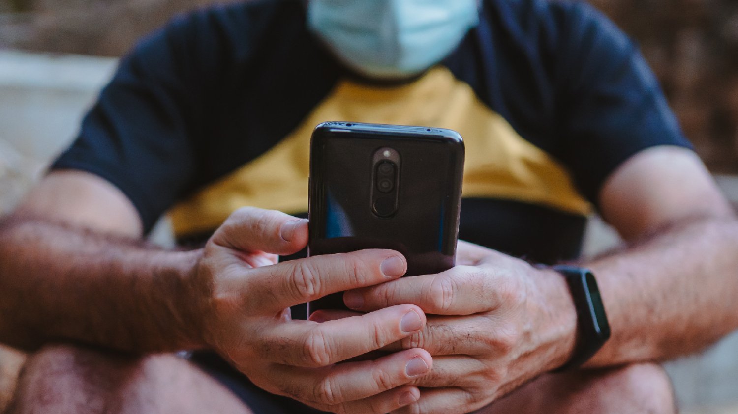 mężczyzna w maseczce siedzący na schodach z telefonem i mający na ręku opaskę
