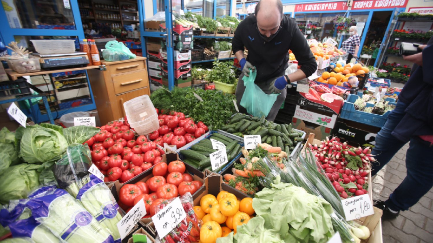 Mężczyzna pakuje produkty wystawione na bazarku z warzywami. Widoczne są między innymi ogórki i pomidory oraz plakietki z ich ceną.