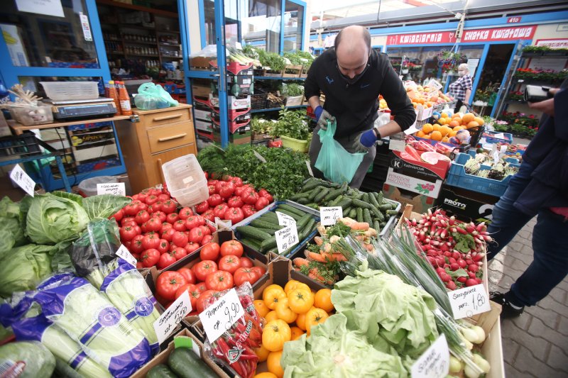 Mężczyzna pakuje produkty wystawione na bazarku z warzywami. Widoczne są między innymi ogórki i pomidory oraz plakietki z ich ceną.