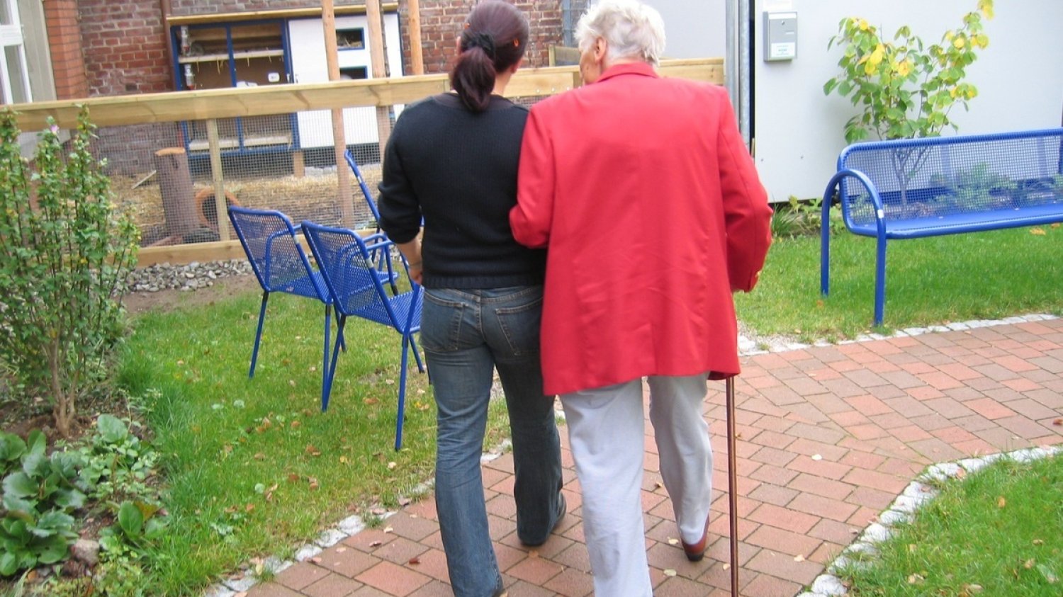 Kobieta prowadzi pod rękę drugą kobietę, w wieku senioralnym.