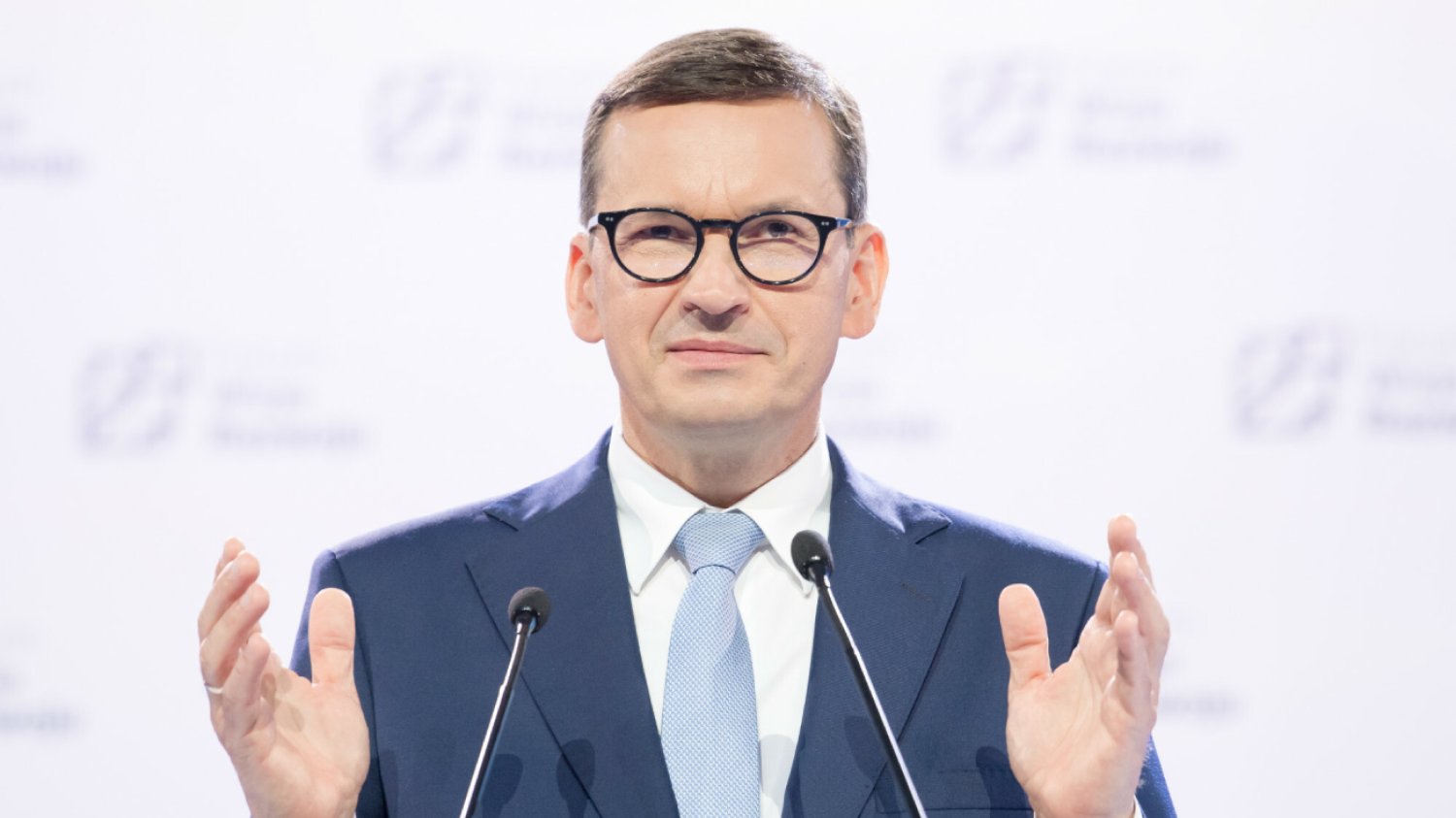 szef rządu Mateusz Morawiecki stoi na białym tle z rozłożonymi rękami na konferencji prasowej