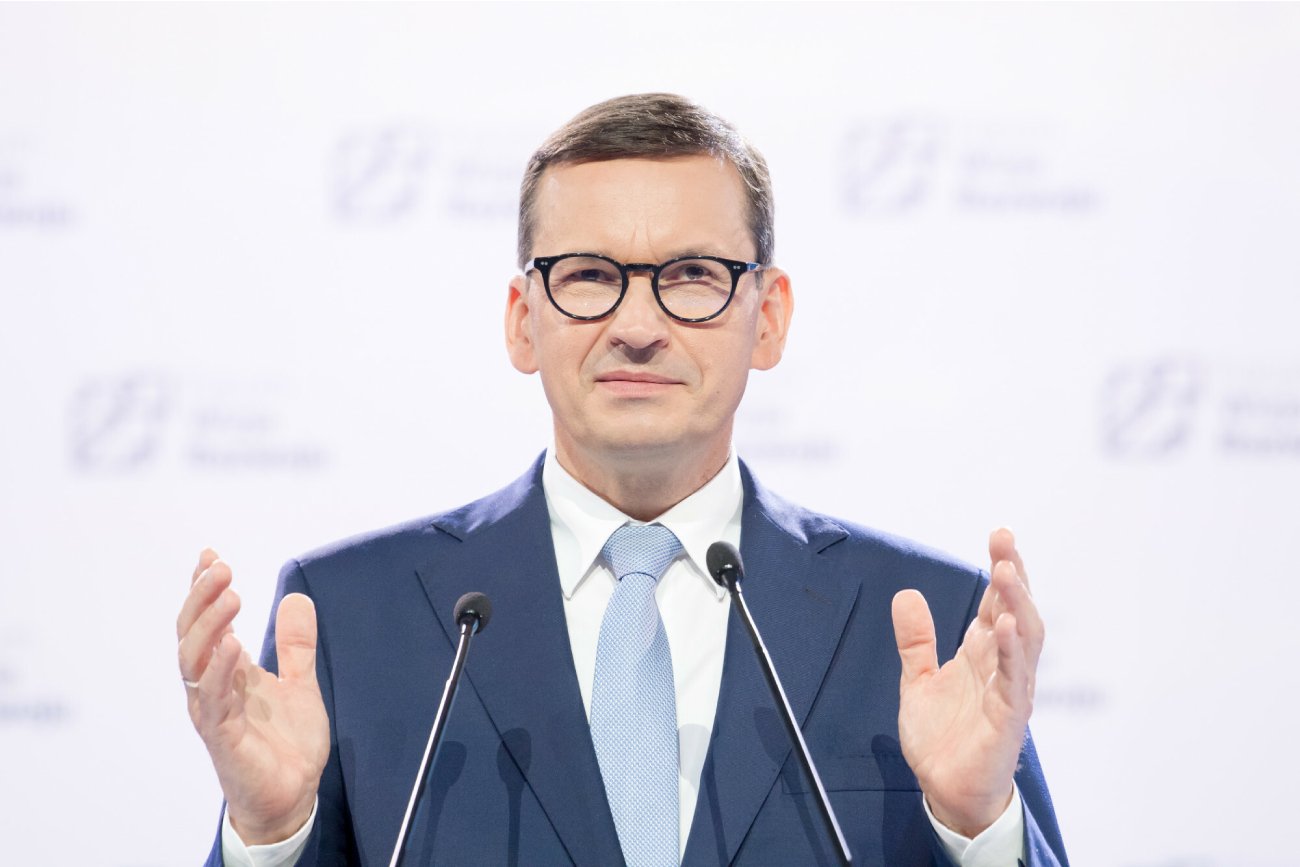 szef rządu Mateusz Morawiecki stoi na białym tle z rozłożonymi rękami na konferencji prasowej