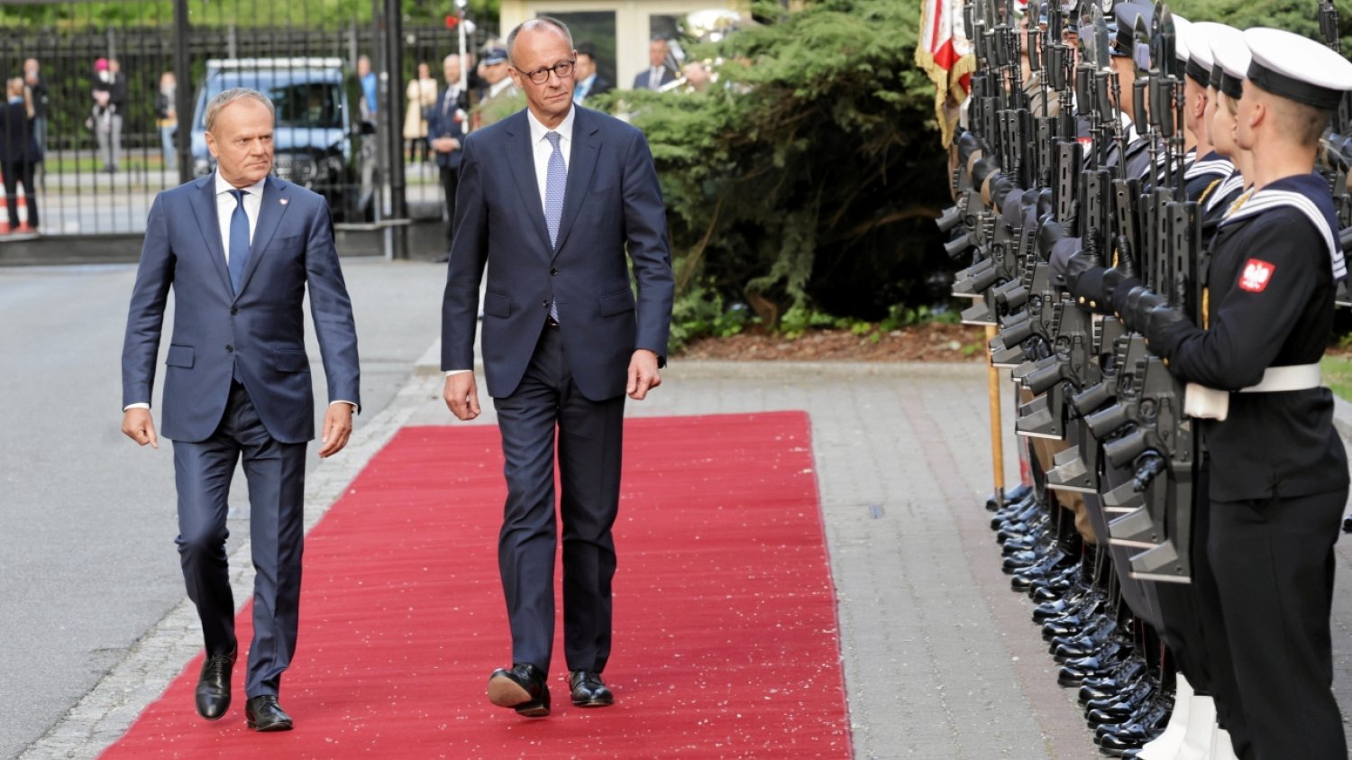 Kanclerz Niemiec Friedrich Merz idący czerwonym dywanem obok premiera Polski Donalda Tuska. Politycy idą wzdłuż szpaleru żołnierzy stojących na baczność.