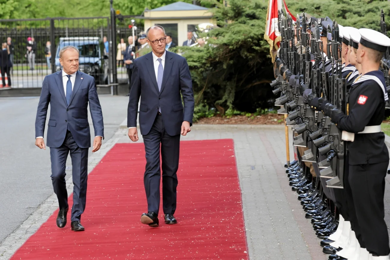 Kanclerz Niemiec Friedrich Merz idący czerwonym dywanem obok premiera Polski Donalda Tuska. Politycy idą wzdłuż szpaleru żołnierzy stojących na baczność.