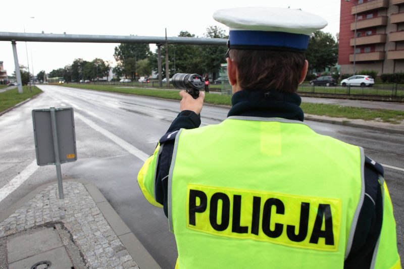 Policjant w jaskrawej kamizelce z napisem "policja" stoi przy miejskiej drodze, w ręku trzyma urządzenie do pomiaru prędkości.