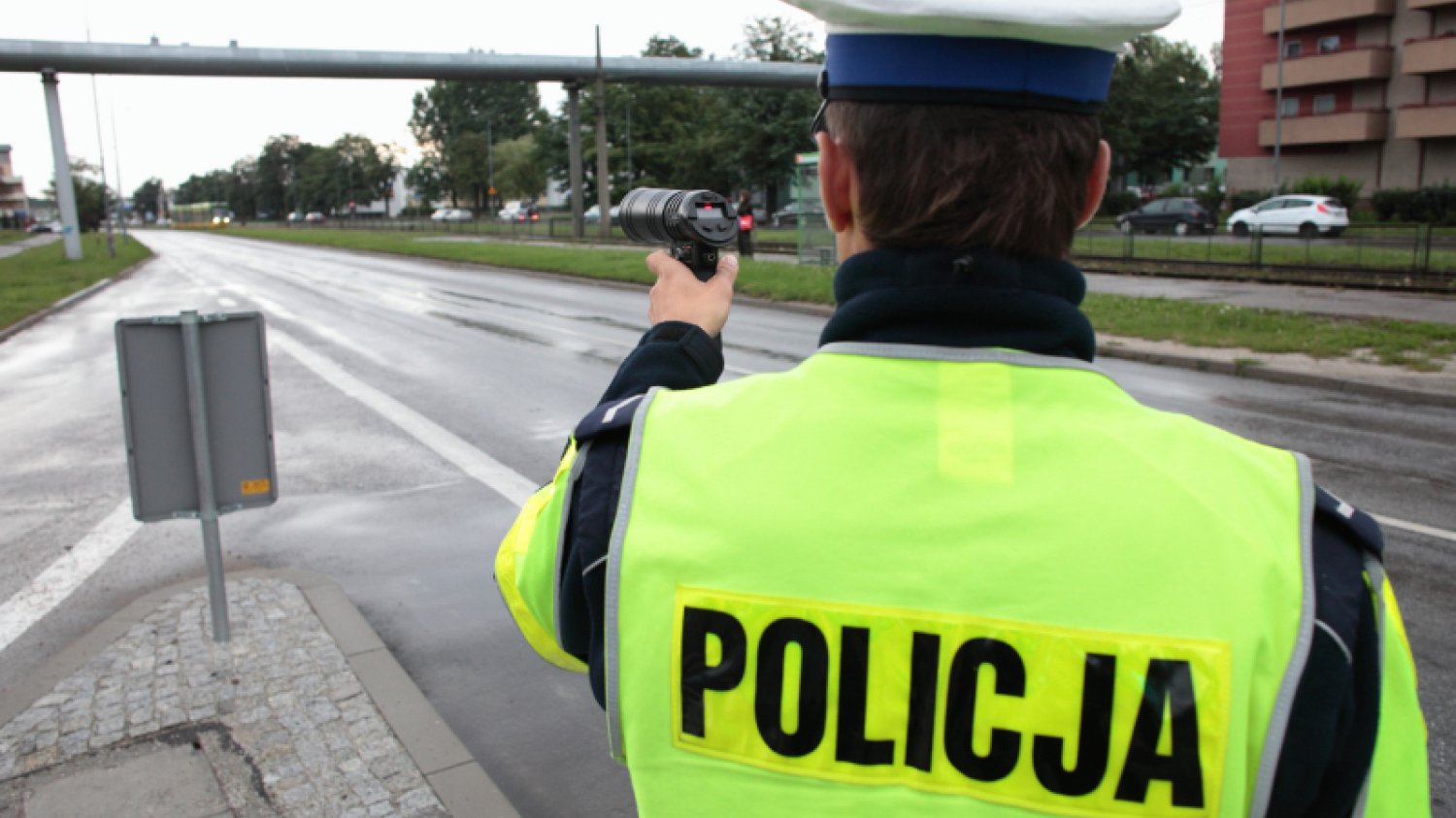 Policjant w jaskrawej kamizelce z napisem "policja" stoi przy miejskiej drodze, w ręku trzyma urządzenie do pomiaru prędkości.