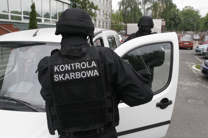 Dwóch mężczyzn ubranych na czarno stojących przy białym samochodzie. Jeden z nich ma na plecach napis "kontrola skarbowa".