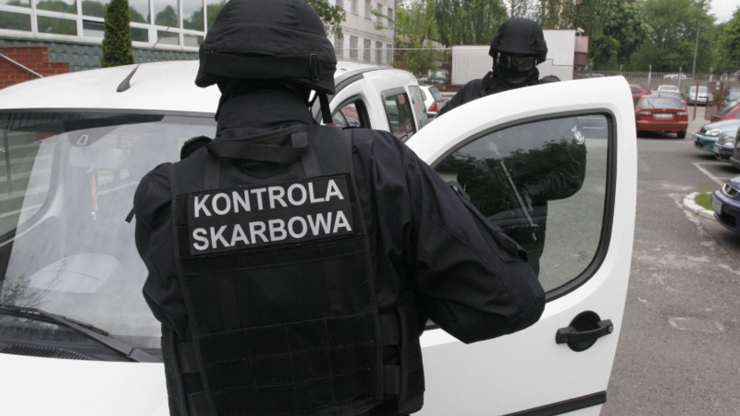Dwóch mężczyzn ubranych na czarno stojących przy białym samochodzie. Jeden z nich ma na plecach napis "kontrola skarbowa".