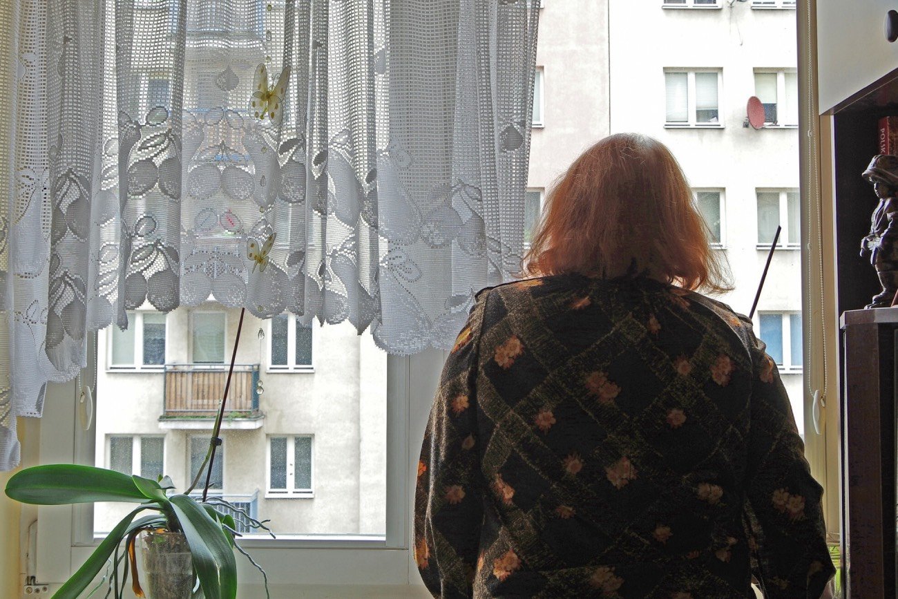 Starsza kobieta stojąca w oknie swojego mieszkania.