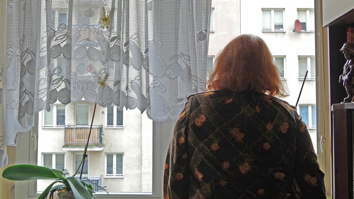 Starsza kobieta stojąca w oknie swojego mieszkania.