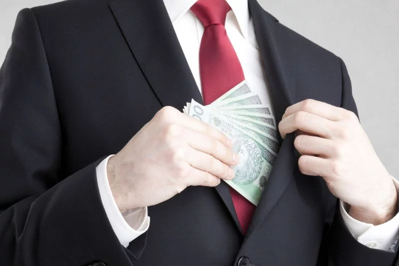 Mężczyzna w garniturze chowa polskie banknoty stuzłotowe do kieszeni marynarki.