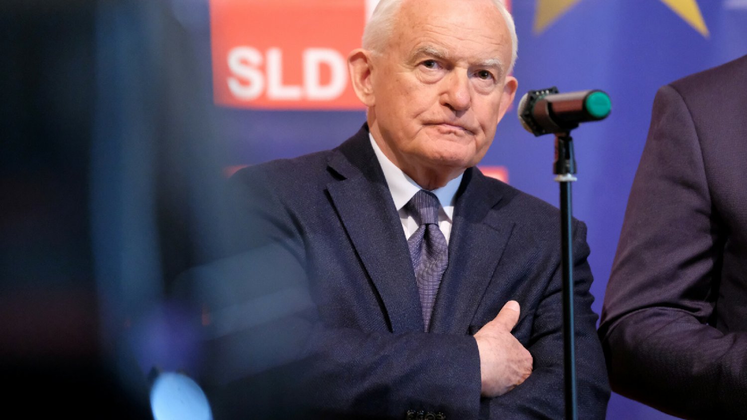 Leczek Miller na konferencji stoi elegancko ubrany przed mikrofonem
