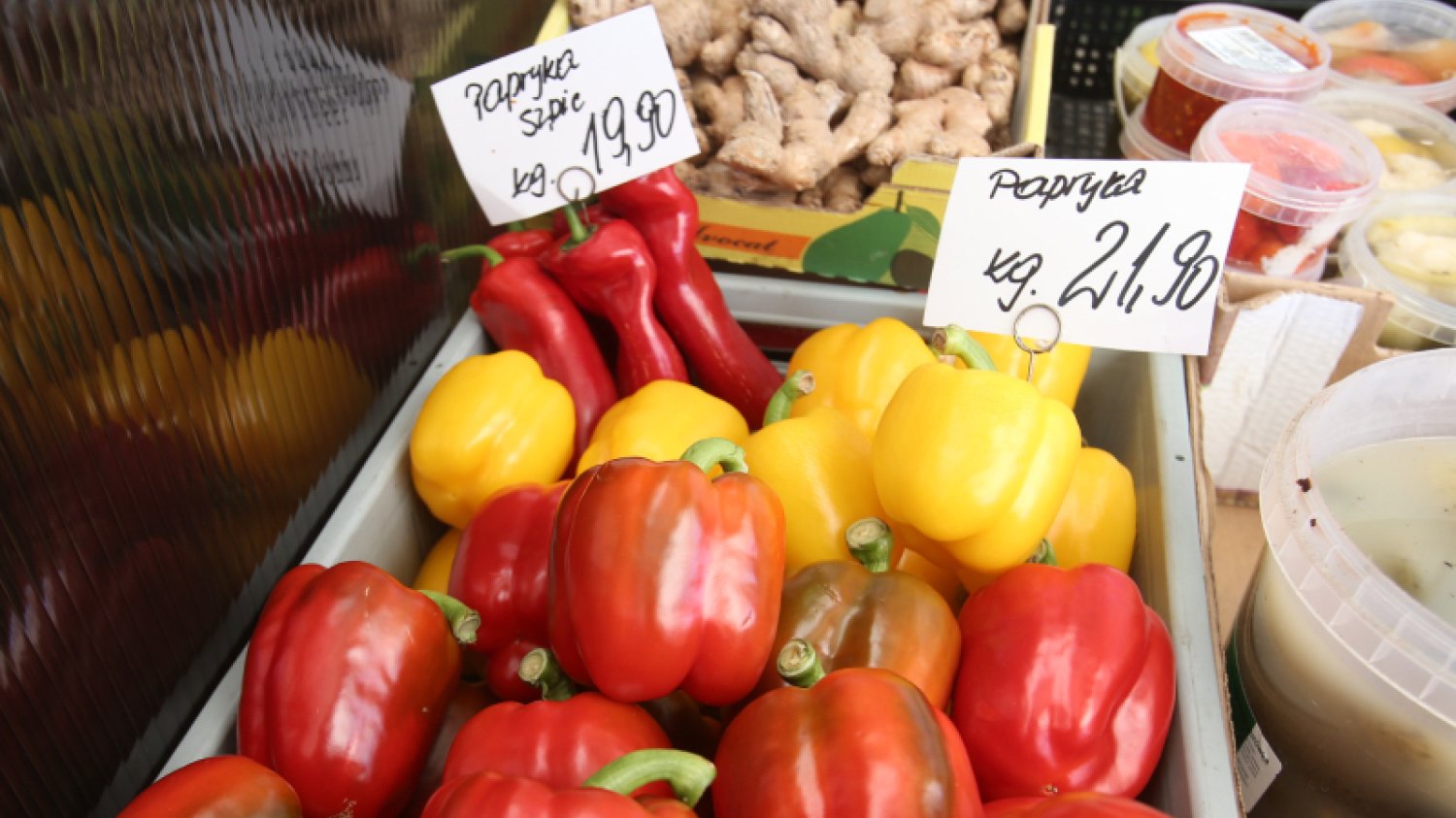 Warzywa wyłożone na stoisku na rynku. Na pierwszym planie papryki z kartkami, na których wypisana jest cena za kilogram.