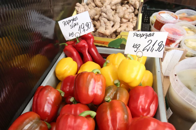Warzywa wyłożone na stoisku na rynku. Na pierwszym planie papryki z kartkami, na których wypisana jest cena za kilogram.