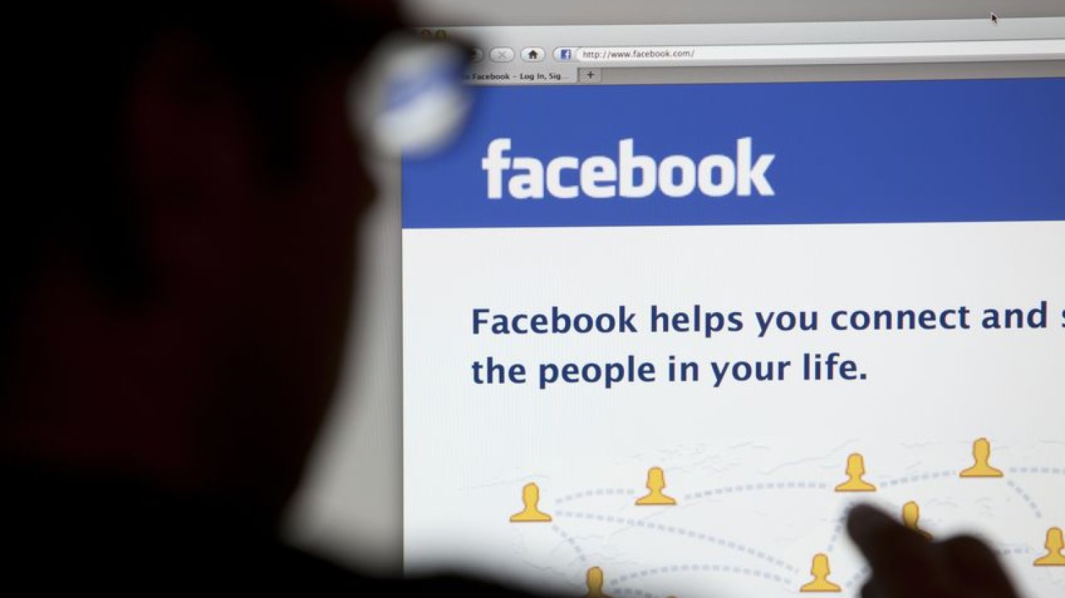 Sylwetka człowieka w okularach który siedzi przed komputerem i loguje się do Facebooka.