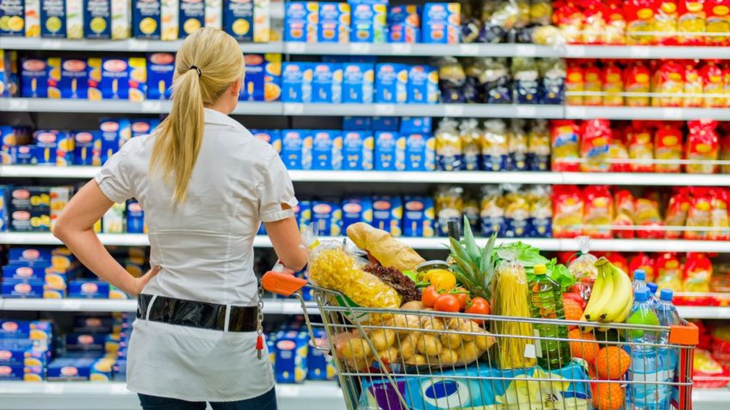 Kobieta stoi z wózkiem zakupowym przed półką w supermarkecie i zastanawia się co wybrać.