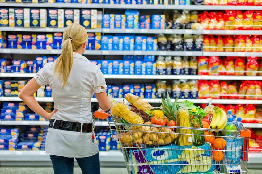 Kobieta stoi z wózkiem zakupowym przed półką w supermarkecie i zastanawia się co wybrać.