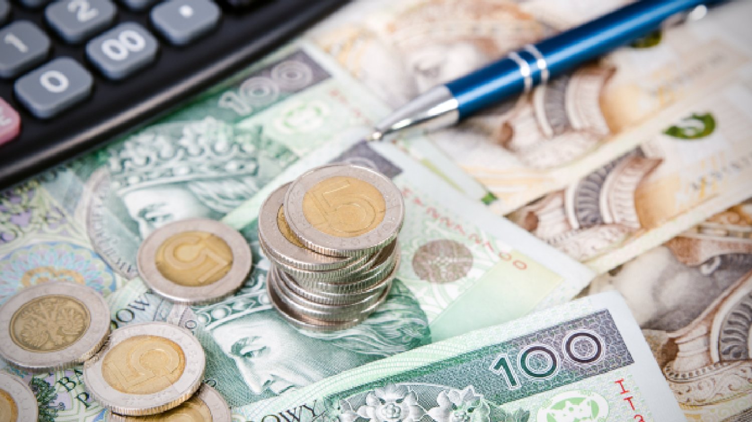 polskie monety leżą na stosie obok banknotów złotowych na tle czarnego kalkulatora i obok niebieskiego długopisu