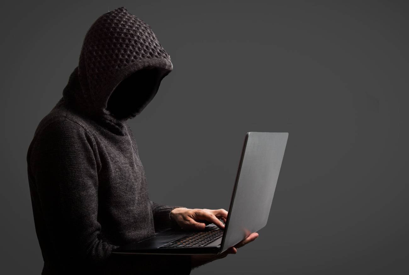 Mężczyzna w kapturze, trzyma w ręku laptop. Nie widać jego twarzy.