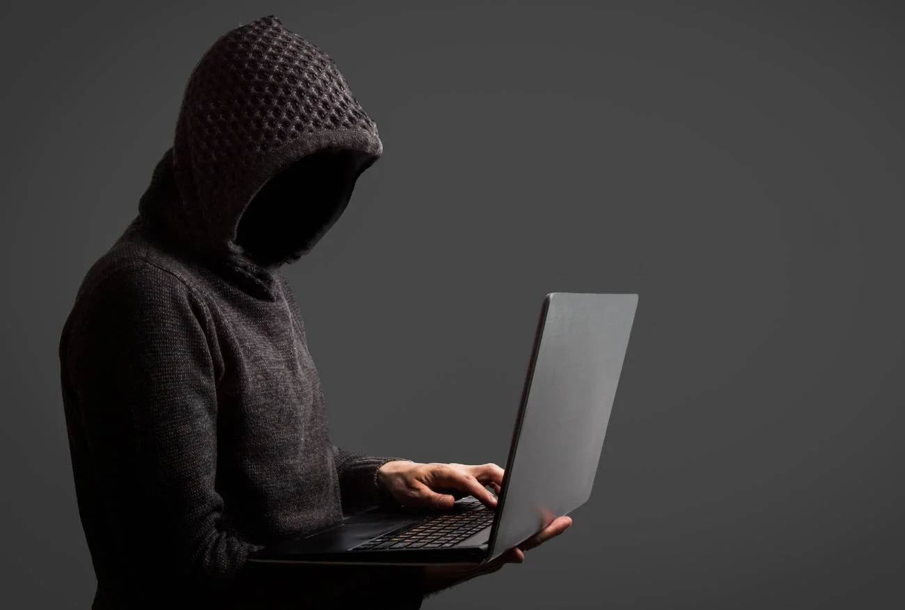 Mężczyzna w kapturze, trzyma w ręku laptop. Nie widać jego twarzy.