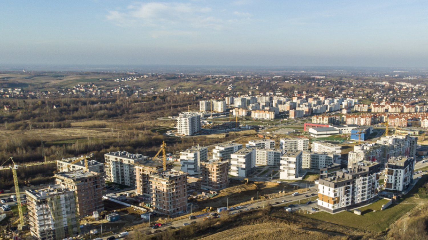 Widok panoramiczny na osiedle mieszkaniowe w Rzeszowie. Na pierwszym planie znajduje się nowe osiedle: część bloków mieszkalnych jest już gotowa, część jest jeszcze w budowie, ustawione przy nich są żurawie.