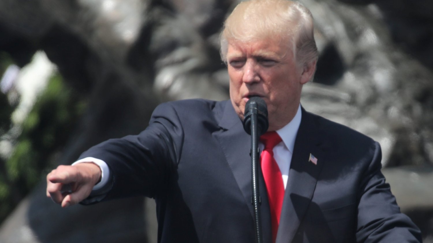 Donald Trump (zdjęcie archiwalne z wizyty w Polsce w 2017 roku). Donald Trumo w garniturze przy mównicy i mikrofonie. Wyciąga przed siebie prawą rękę i wskazuje na coś wyprostowanym palcem wskazującym.