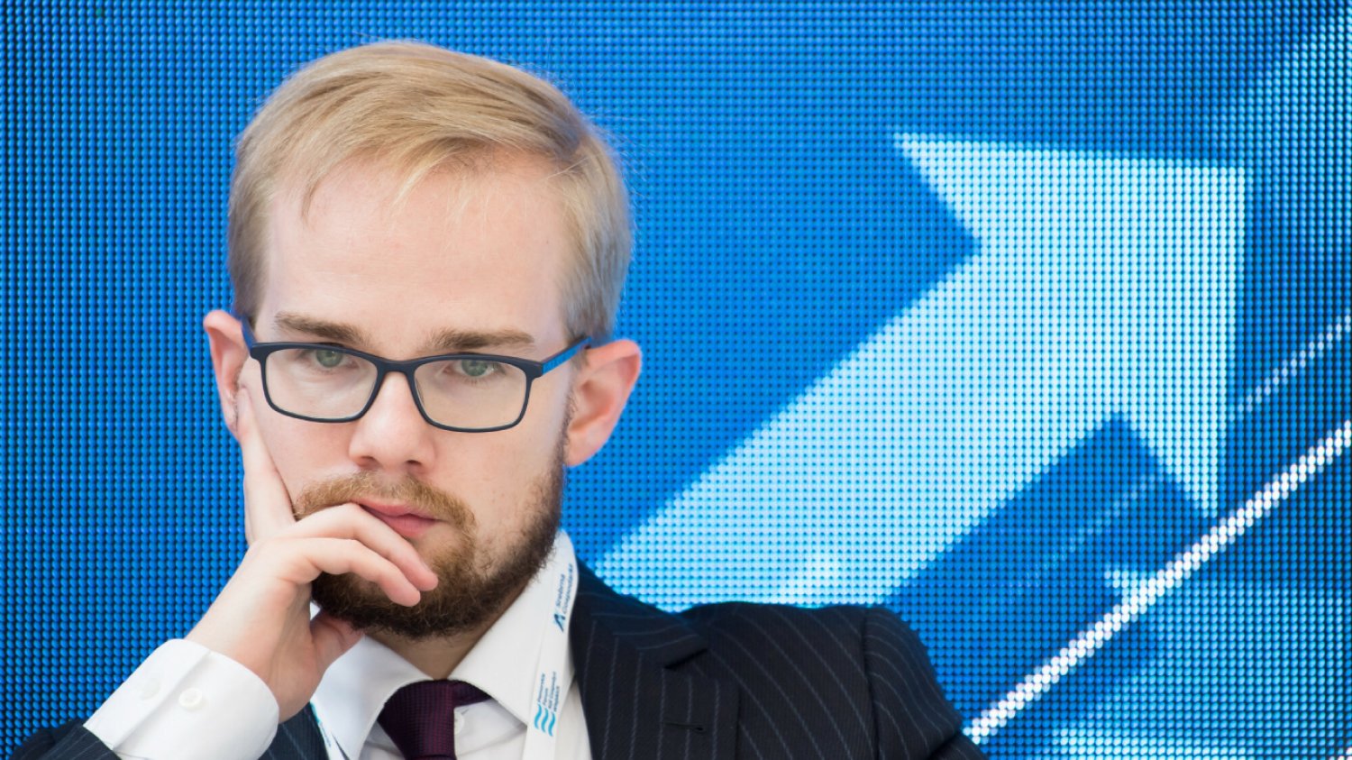 zamyślony wiceminister finansów Piotr Patkowski siedzi w okularach na niebieskim tle