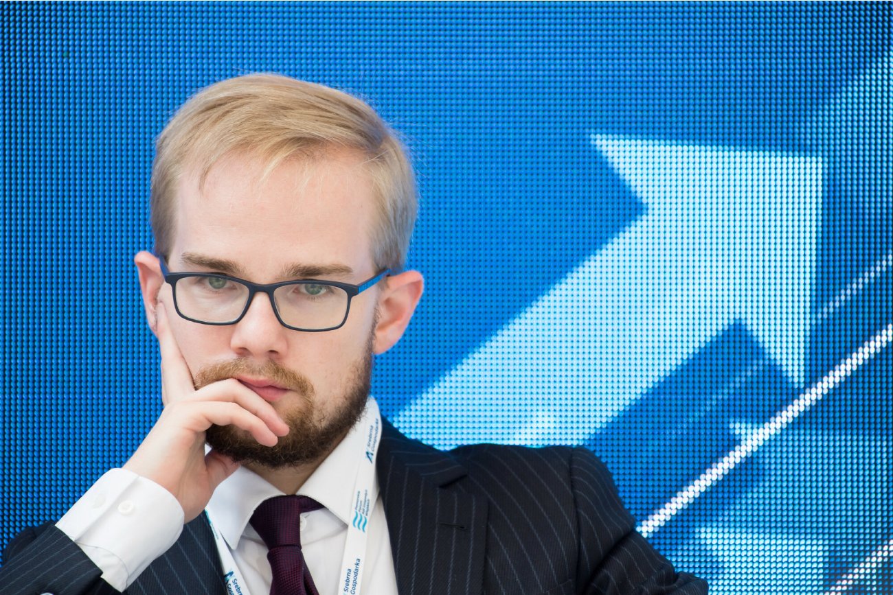 zamyślony wiceminister finansów Piotr Patkowski siedzi w okularach na niebieskim tle