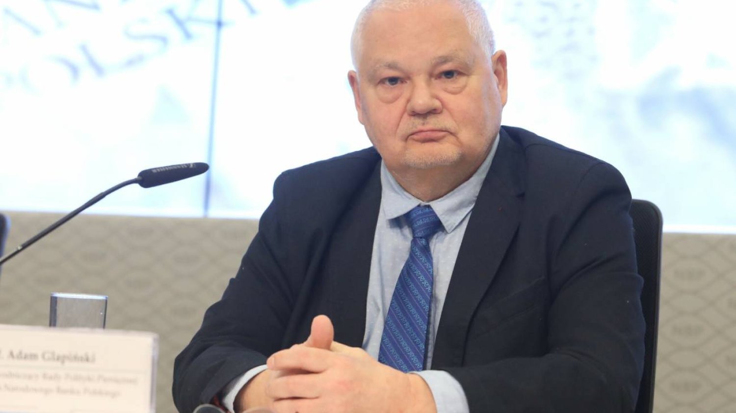 prezes NBP Adam Glapiński w niebieskim garniturze patrzy prosto w obiektyw fotografa na konferencji prasowej