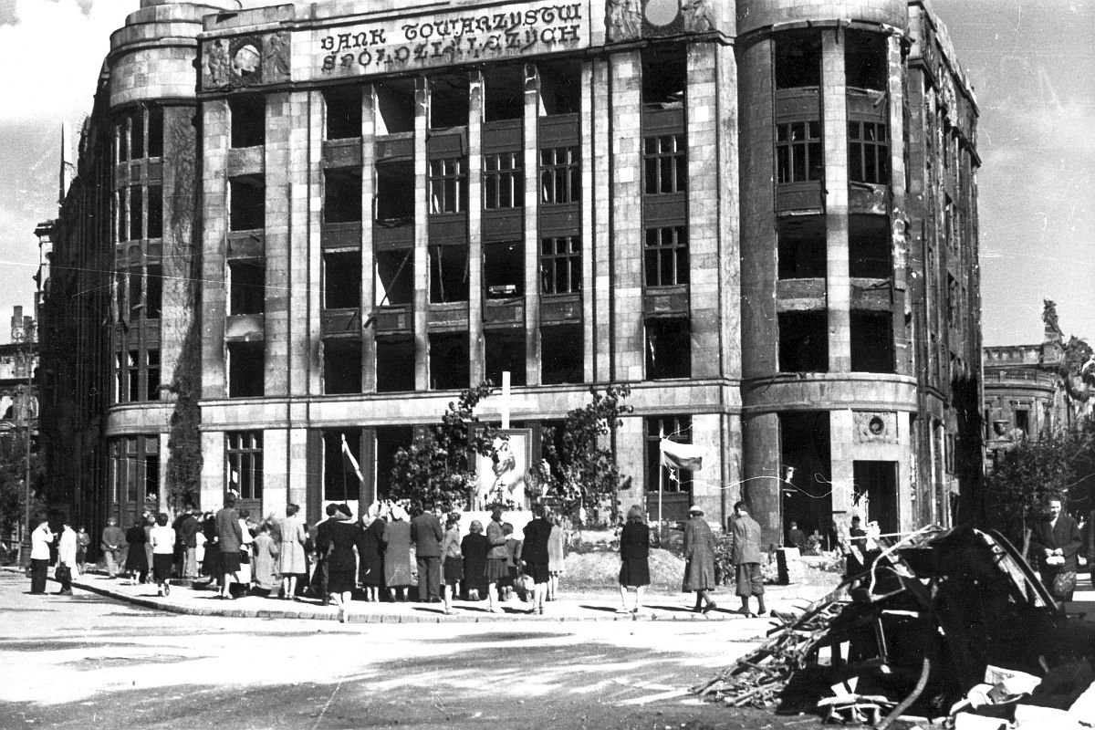 Pierwsza rocznica powstania warszawskiego - ludność zgromadzona wokół ołtarza ulicznego ustawionego przed uszkodzonym gmachem Banku Towarzystw Spółdzielczych (tzw. Dom "Pod Orłami" ul. Jasna 1 róg ul. Zgoda). Widoczny fragment budynku filharmonii