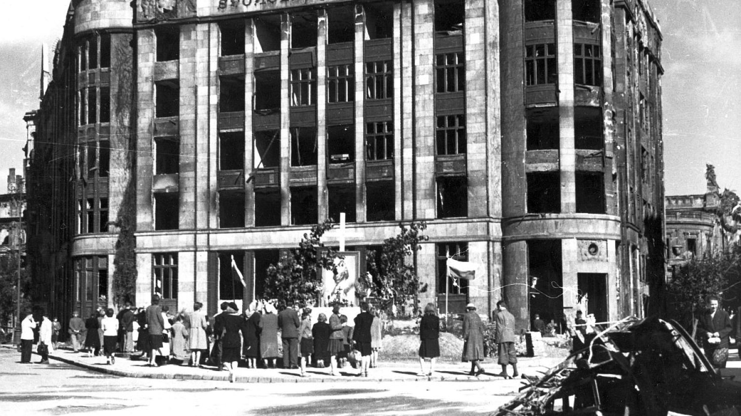 Pierwsza rocznica powstania warszawskiego - ludność zgromadzona wokół ołtarza ulicznego ustawionego przed uszkodzonym gmachem Banku Towarzystw Spółdzielczych (tzw. Dom "Pod Orłami" ul. Jasna 1 róg ul. Zgoda). Widoczny fragment budynku filharmonii