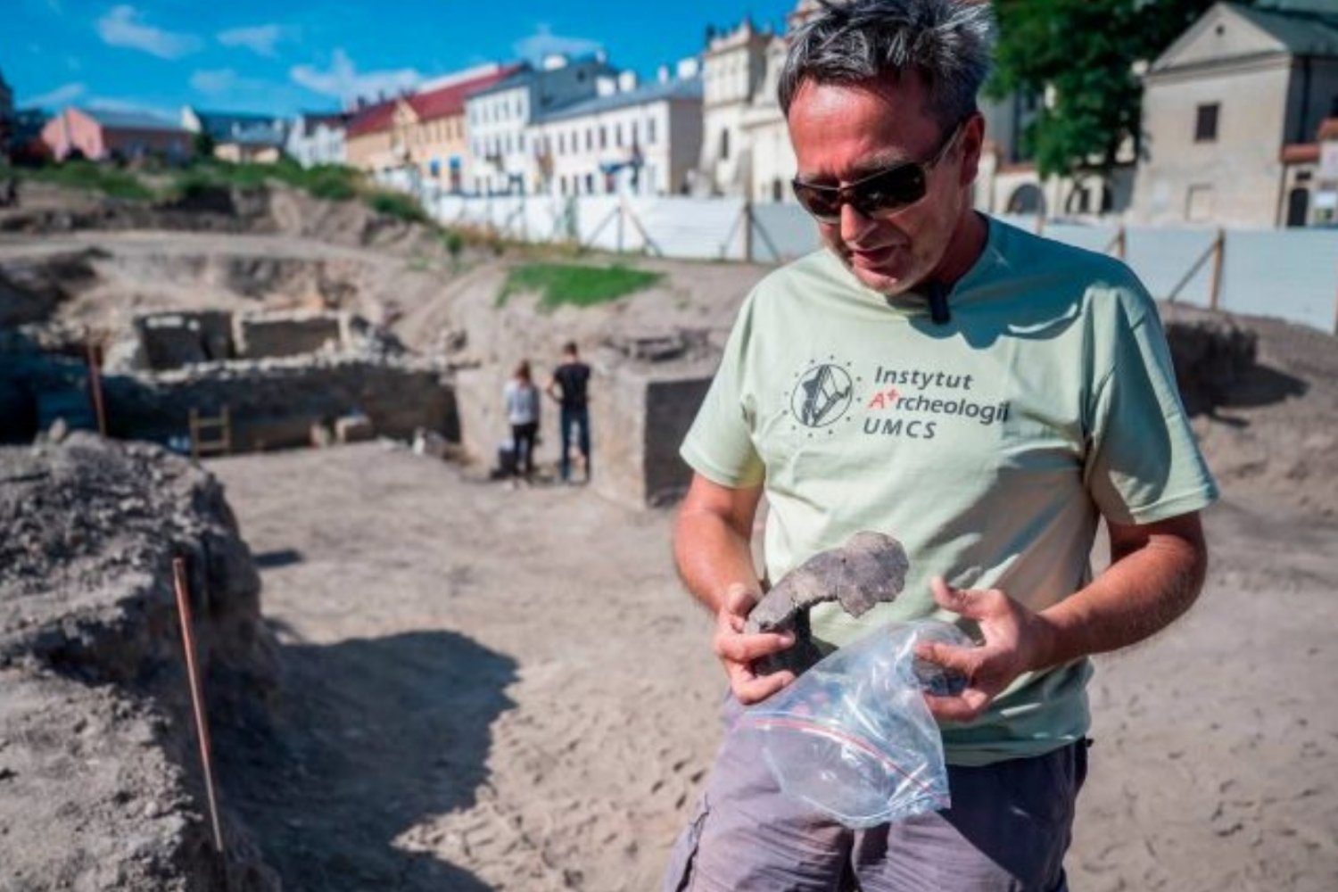 Archeolog z jednym ze znalezisk w ręku na pierwszym planie. Na drugim planie pozostali archeolodzy podczas prac.