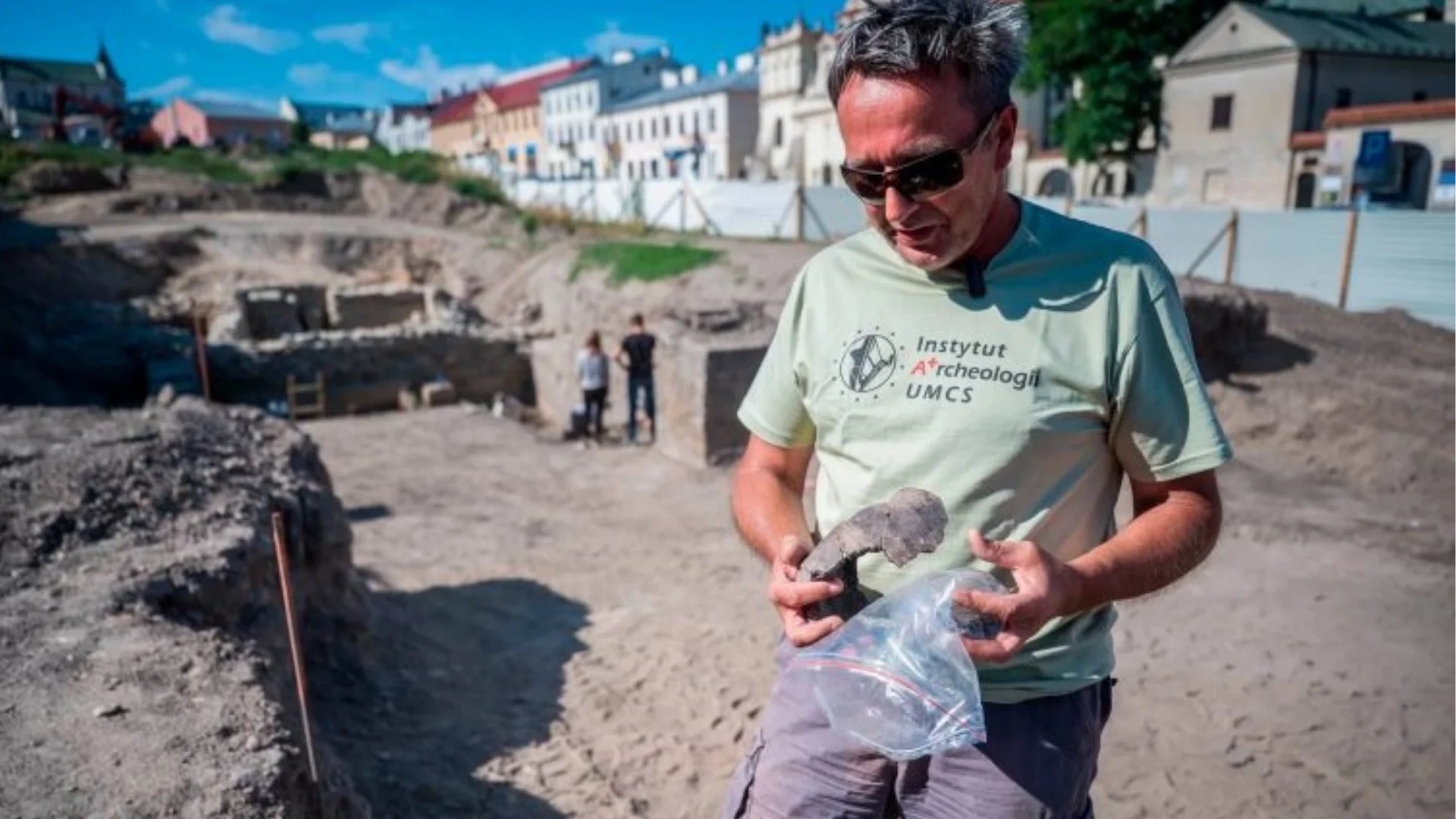 Archeolog z jednym ze znalezisk w ręku na pierwszym planie. Na drugim planie pozostali archeolodzy podczas prac.