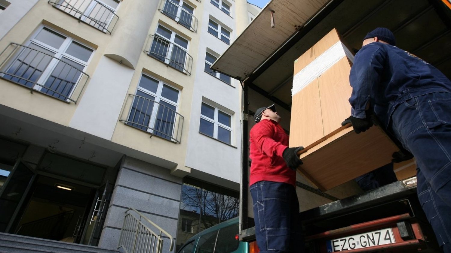 Dwóch mężczyzn z firmy przeprowadzkowej wyciąga z ciężarówki szafę. Zaraz wniosą ją do żółtego budynku stojącego obok