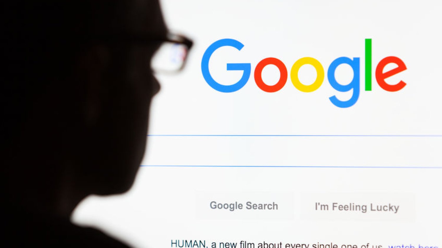 Ciemna sylwetka mężczyzny siedzącego przed monitorem z wyświetloną przeglądarką Google.