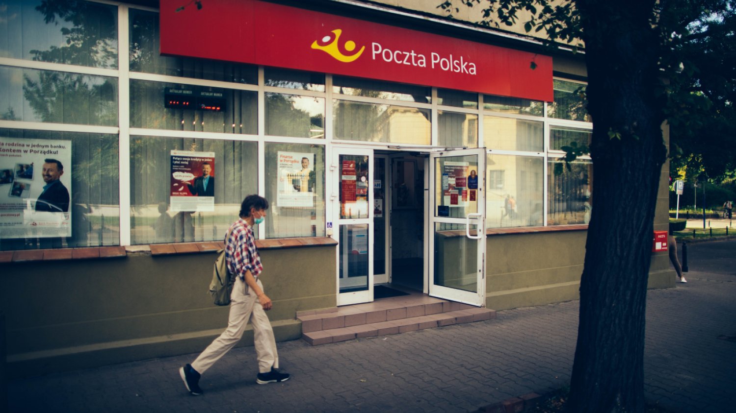 Mężczyzna w letnim ubraniu przechodzący koło otwartej placówki Poczty Polskiej.
