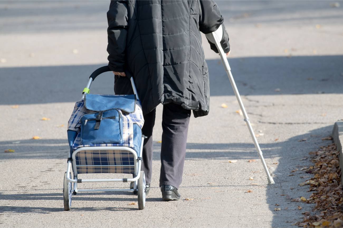 Emerytka idzie chodnikiem z torbą na kółkach, opierając się o kulę