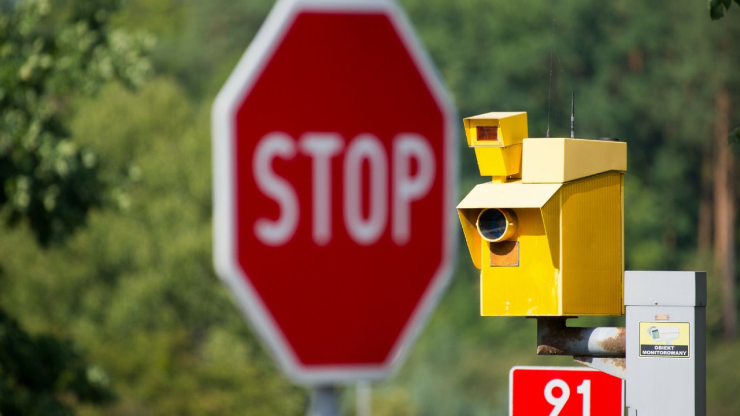 żółty fotoradar na tle lasu i czerwonego znaku stop