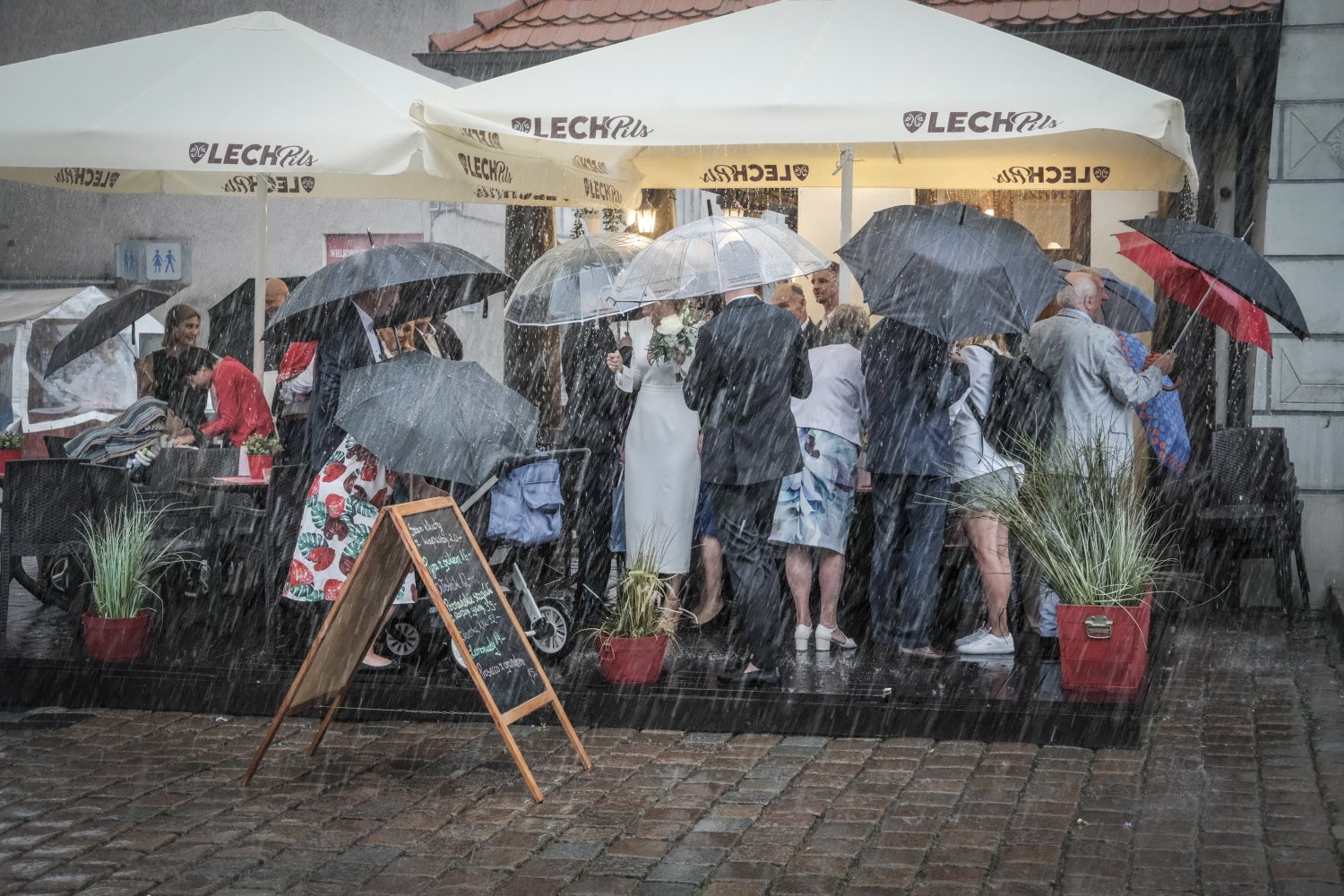 Ludzie z parasolkami chowają się pod parasolem ogrodowym w ulewny dzień.